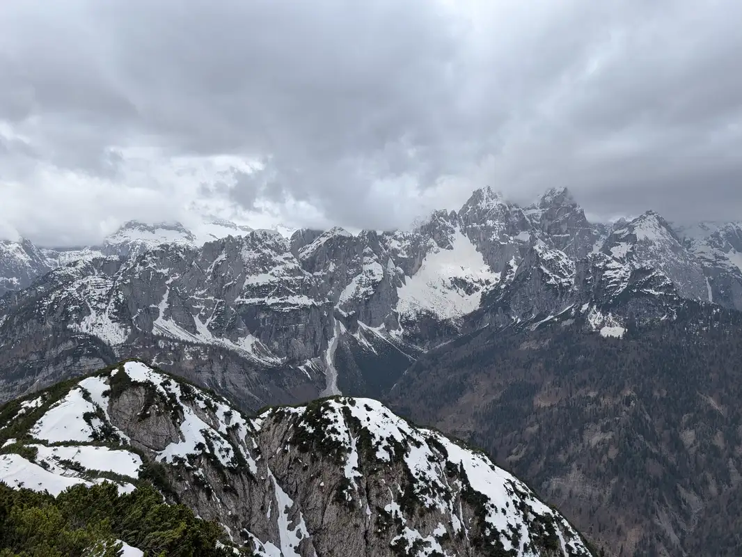 View from the top of Monte Re into southern direction, in the foreground on the left the pre-summit is visible. In the background we can see Jôf Fuart group with its peaks hidden under the clouds. Top of Monte Re is flat with lots of spots to sit, there is also an interesting hiking signpost referring to the mining traditions.